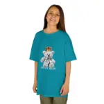 Young girl smiling while wearing an oversized teal blue t-shirt featuring a teddy bear wearing a crown graphic with 'TEDDY BEAR' text, paired with olive green pants on a white background