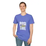 Man smiling at camera wearing periwinkle blue t-shirt with 'POSITIVE VIBES' graphic in white and gold text, casual pose with hand in jeans pocket against white background